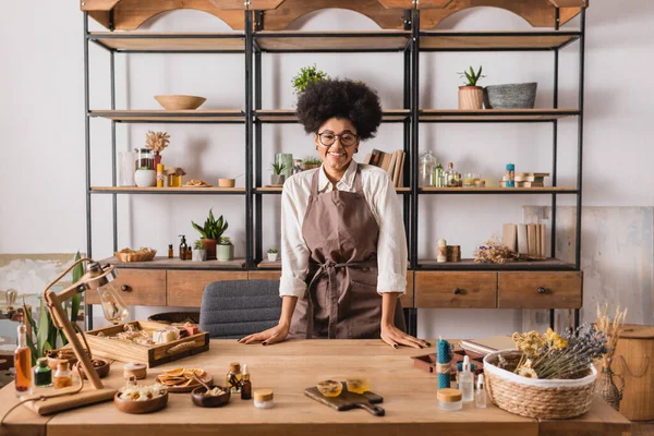 Mujer afroamericana feliz en delantal de pie a la mesa con jabón hecho a mano y velas cerca de ingredientes naturales en el taller - foto de stock