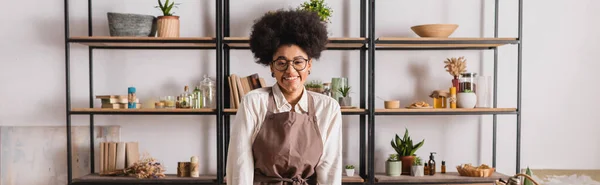 Mujer afroamericana feliz en gafas y delantal de pie cerca de estante borroso con libros e ingredientes naturales en taller de artesanía, pancarta - foto de stock
