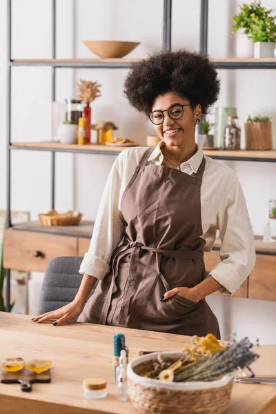 Happy african american woman with hand in pocket of apron standing near table with blurred soap bars and dried herbs - foto de stock