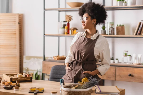 Mulher afro-americana com as mãos em bolsos de avental de pé perto de sabão artesanal e ingredientes em oficina de artesanato — Fotografia de Stock