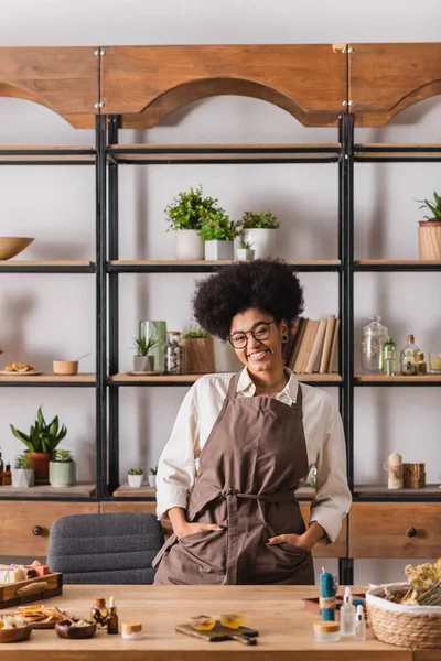 Sorrindo mulher afro-americana de pé com as mãos em bolsos de avental perto de ingredientes naturais para fabricação de sabão e vela — Fotografia de Stock