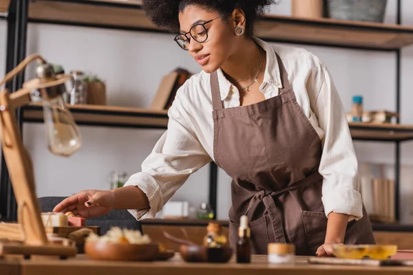 African american woman in apron and eyeglasses taking soap bars near natural ingredients on blurred foreground — Stock Photo