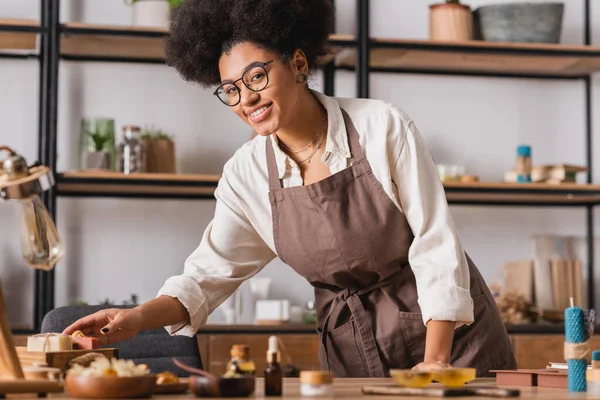 Sonriente artesana afroamericana en gafas y delantal llegando a jabón hecho a mano cerca de ingredientes borrosos en la mesa - foto de stock