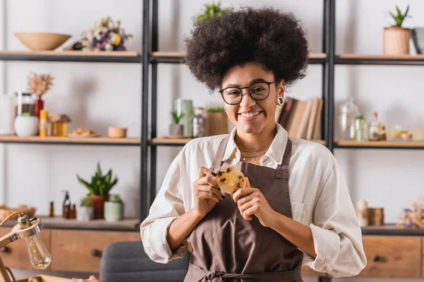 Alegre mujer afroamericana en gafas y delantal sosteniendo jabón hecho a mano cerca de rack con ingredientes naturales sobre fondo borroso - foto de stock