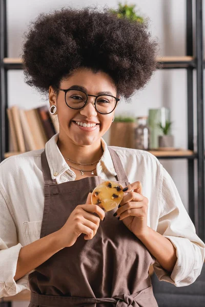 Joyful african american craftswoman in eyeglasses and apron holding bar of herbal soap in blurred workshop — Stock Photo