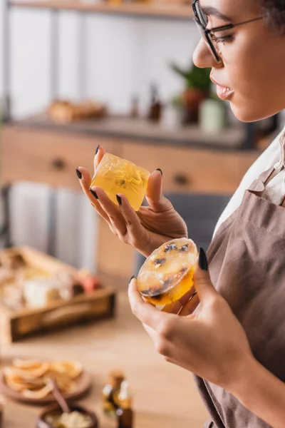 African american craftswoman in eyeglasses and apron holding herbal soap in blurred workshop — Foto stock