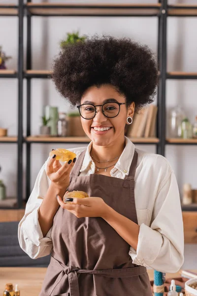 Cheerful african american woman holding bars of handmade soap and smiling at camera in craft workshop — Foto stock