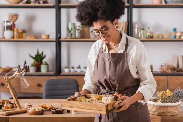Pleased african american craftswoman in eyeglasses and apron holding wooden tray with handmade soap in workshop — Stockfoto