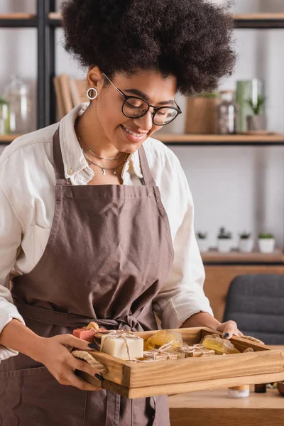 Donna afroamericana sorridente in grembiule e occhiali vassoio con assortimento di sapone fatto in casa in laboratorio artigianale — Foto stock