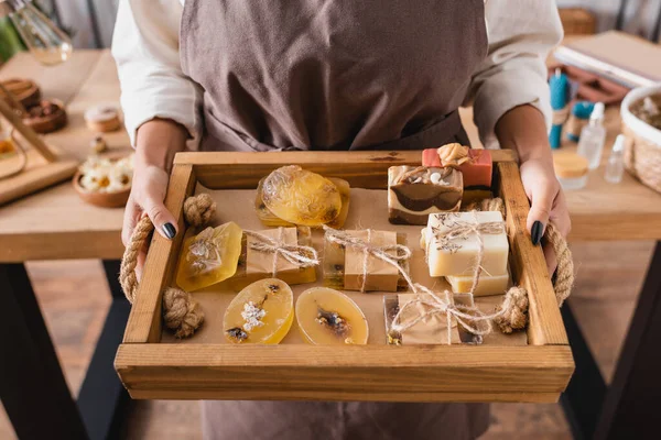 Wooden tray with assortment of handmade soap in hands of cropped african american craftswoman - foto de stock