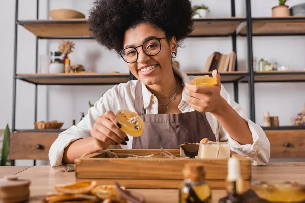 Happy african american craftswoman in eyeglasses holding handmade soap near natural ingredients on blurred foreground — Fotografia de Stock