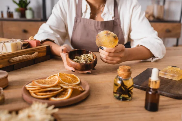 Partial view of african american woman holding homemade soap and wooden bowl with dried herbs near dried orange slices and essential oils — Stock Photo