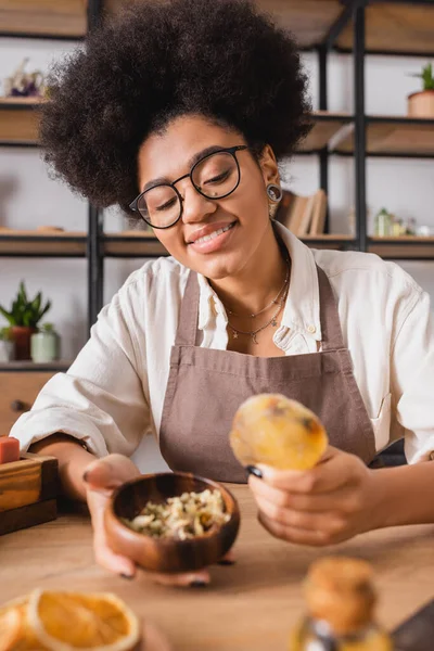 Happy african american craftswoman holding natural homemade soap and wooden bowl with dried herbs in workshop — Stock Photo