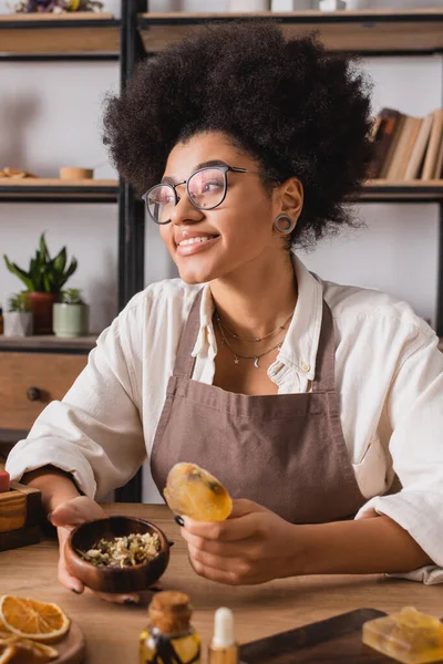 Smiling african american woman holding handmade soap and wooden bowl with dried herbs in craft workshop — Stock Photo