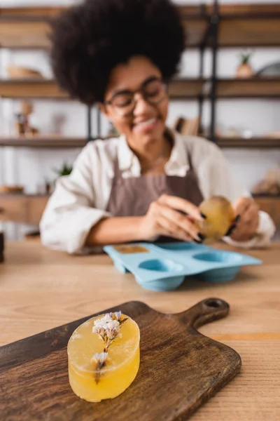 Foco seletivo de sabão à base de plantas na tábua de corte perto de artesã afro-americana sorrindo no fundo borrado — Fotografia de Stock