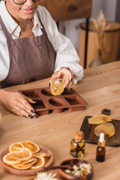 Vista parcial de la artesana afroamericana con jabón hecho a mano cerca del molde de silicona y los ingredientes naturales en la mesa de madera - foto de stock