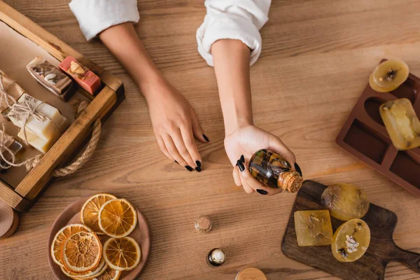 Top view of cropped african american woman holding essential oil near homemade soap and dried orange slices in craft workshop - foto de stock
