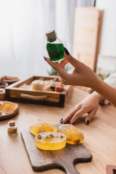 Cropped view of african american woman holding jar with essential oil near handmade soap on chopping board - foto de stock