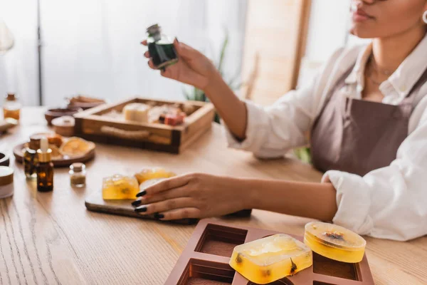 Selective focus of soap bars and silicone mold near cropped african american woman holding essential oil on blurred background — Stock Photo