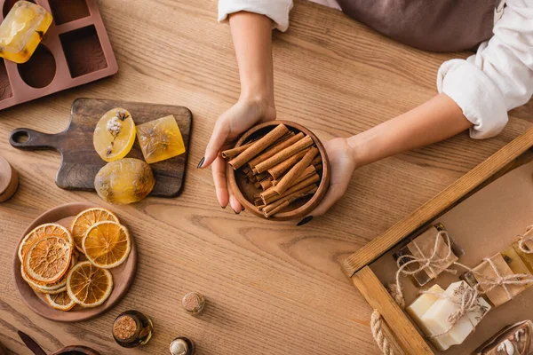 Top view of cropped african american woman holding cinnamon sticks near dried orange slices and handmade soap on wooden table — Stockfoto
