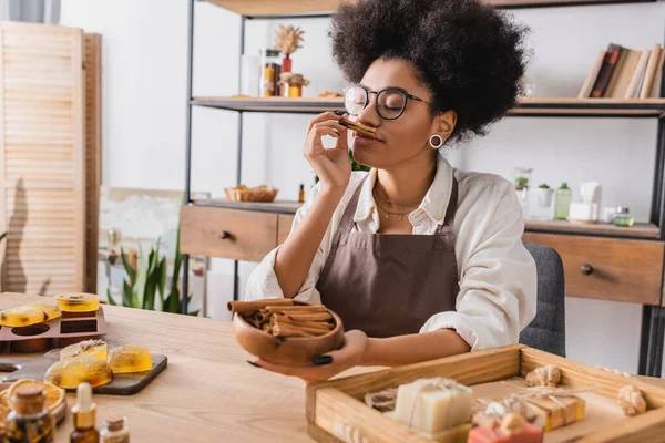 Pleased african american craftswoman in eyeglasses enjoying flavor of cinnamon near handmade soap in workshop — Stockfoto