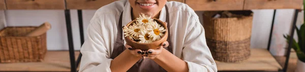 Partial view of happy african american woman holding wooden bowl with dried camomiles in craft workshop, banner — Stock Photo