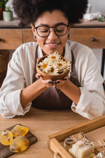 Happy african american craftswoman in eyeglasses holding wooden bowl with dried camomiles near bars of handmade soap — Stock Photo