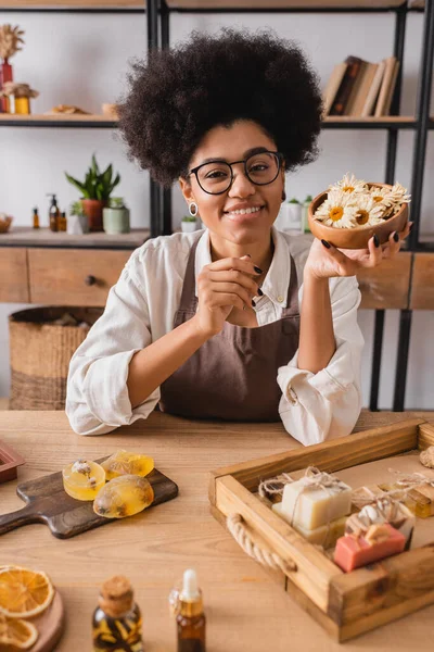 Fröhliche afrikanisch-amerikanische Frau in Brille und Schürze mit getrockneten Kamillen in der Nähe von Seifenstangen und natürlichen Zutaten Bastelwerkstatt — Stockfoto