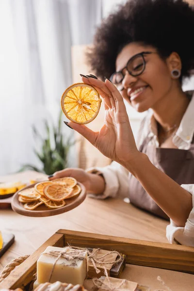 Blurred african american woman in eyeglasses holding dried orange slices near wooden tray with handmade soap — Stock Photo