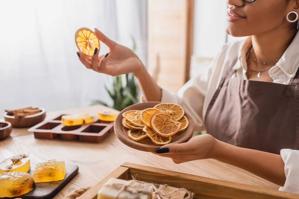 Abgeschnittene Ansicht einer lächelnden Afroamerikanerin mit einem Holzteller mit getrockneten Orangenscheiben in der Nähe handgemachter Seife in der Werkstatt — Stockfoto