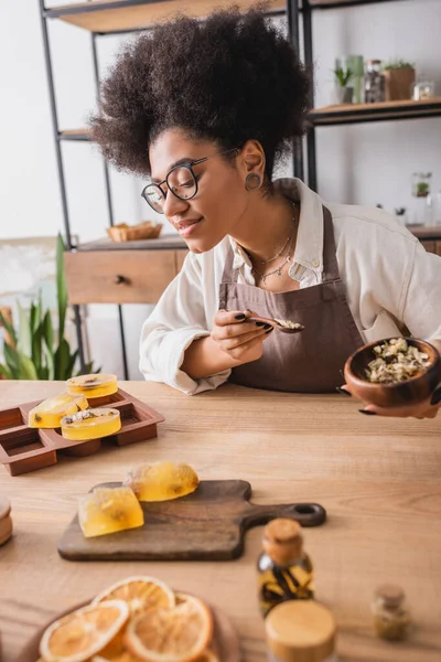 Pleased african american craftswoman in eyeglasses holding dried herbs near soap bars and silicone mold on wooden table — Stock Photo
