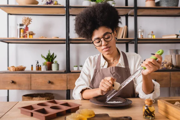 Afro-americana artigiana in occhiali grattugiando sapone bar vicino stampo in silicone e olio essenziale — Foto stock