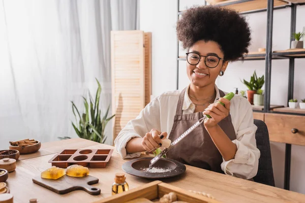 Happy african american craftswoman in eyeglasses looking at camera while grating soap bar near silicone mold in workshop — Stock Photo