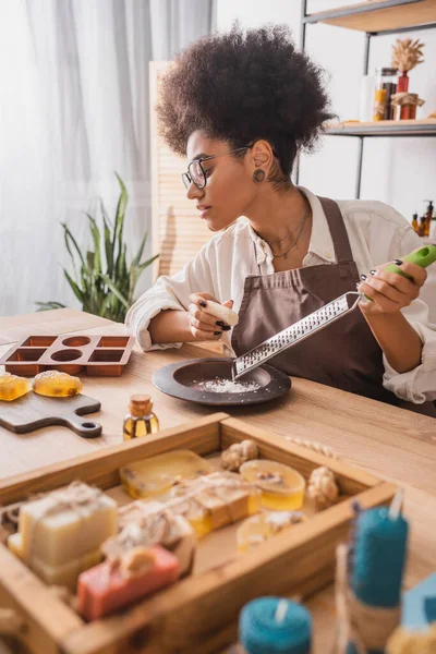 African american woman holding grater near silicone mold and handmade soap in craft workshop - foto de stock