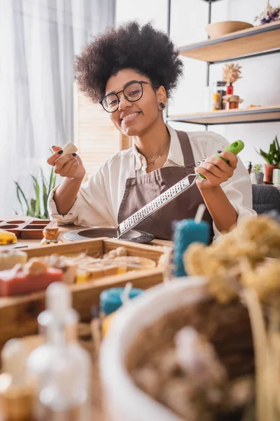 Cheerful african american woman with soap bar and grater looking at camera on blurred foreground in crafts workshop — Foto stock