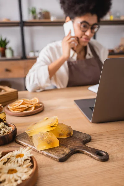 Selective focus of homemade soap near laptop and blurred african american woman accepting order on smartphone — Stock Photo
