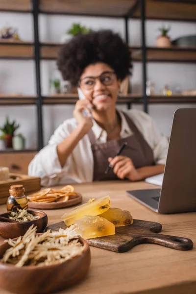 Selective focus of dried herbs and handmade soap near computer and blurred african american woman accepting order on cellphone — Stock Photo