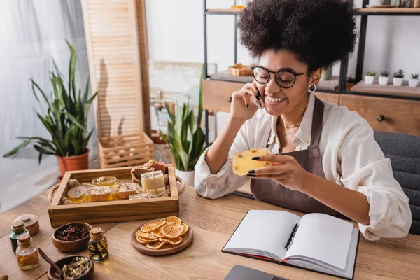 Happy african american woman holding homemade soap and talking on cellphone near natural ingredients and notebook — Stockfoto