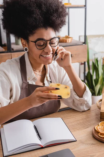 Cheerful african american craftswoman in eyeglasses holding handmade soap while talking on smartphone near notebook — Stock Photo