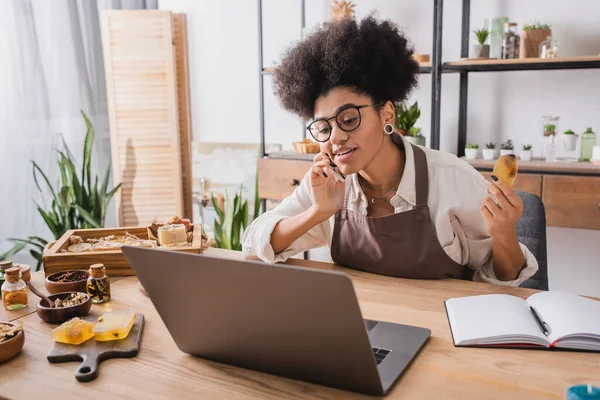 Positive african american craftswoman talking on mobile phone near laptop and homemade soap in workshop — Stockfoto