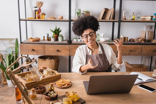 Happy african american artisan in eyeglasses and apron gesturing during video call near handmade soap in workshop — Stockfoto