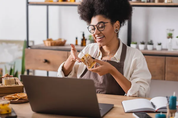 Happy african american woman in eyeglasses showing handmade soap during video call on laptop in craft workshop — Stock Photo