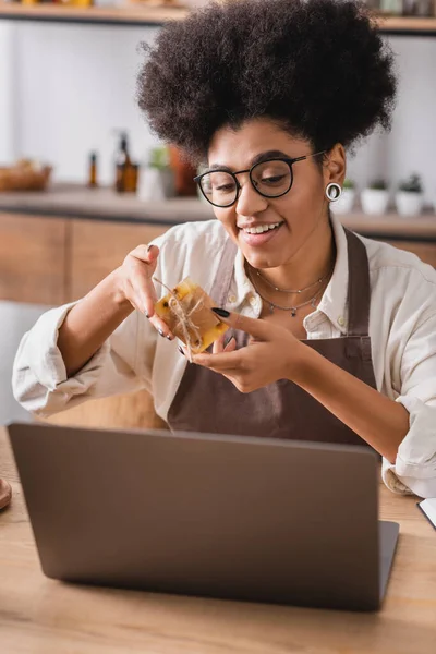 Joyful african american craftswoman in eyeglasses and apron showing homemade soap during video chat on blurred laptop — Stockfoto