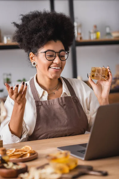 Cheerful african american craftswoman in eyeglasses and apron holding handmade soap during video chat on blurred laptop — Stock Photo