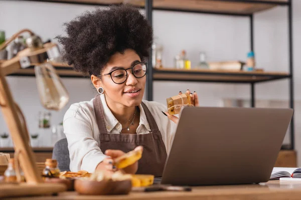 Positive african american craftswoman holding soap bars while having video chat on laptop on blurred foreground — Stockfoto