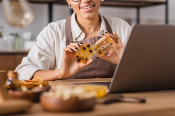 Vista recortada de la sonriente artesana afroamericana mostrando jabón de hierbas mientras tiene videollamada en el portátil - foto de stock
