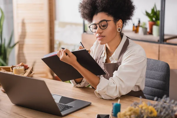 African american craftswoman in apron and eyeglasses writing in notebook near laptop and handmade products on blurred foreground — Stockfoto