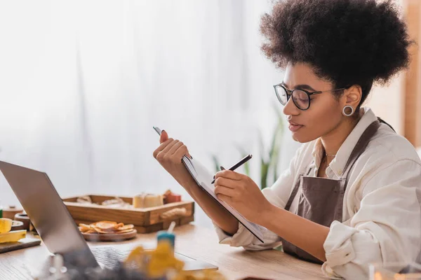 Curly african american woman in eyeglasses writing in notebook near laptop and blurred products in craft workshop — Stock Photo