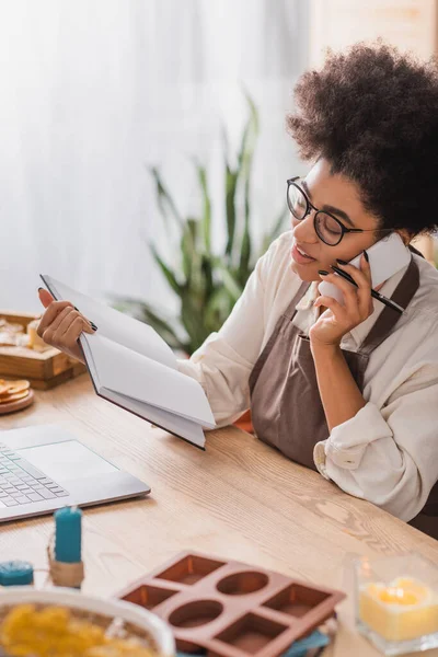 African american woman in apron and eyeglasses holding notebook and talking on cellphone near laptop in craft workshop — Stock Photo