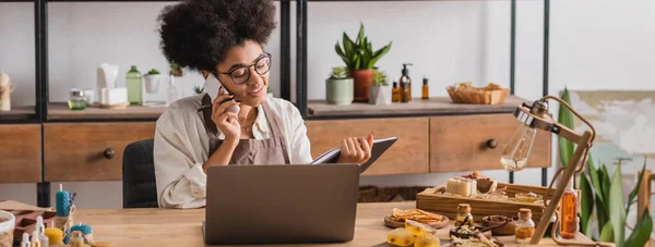 Smiling african american craftswoman looking in notebook and talking on smartphone near laptop and handmade products, banner — Stock Photo